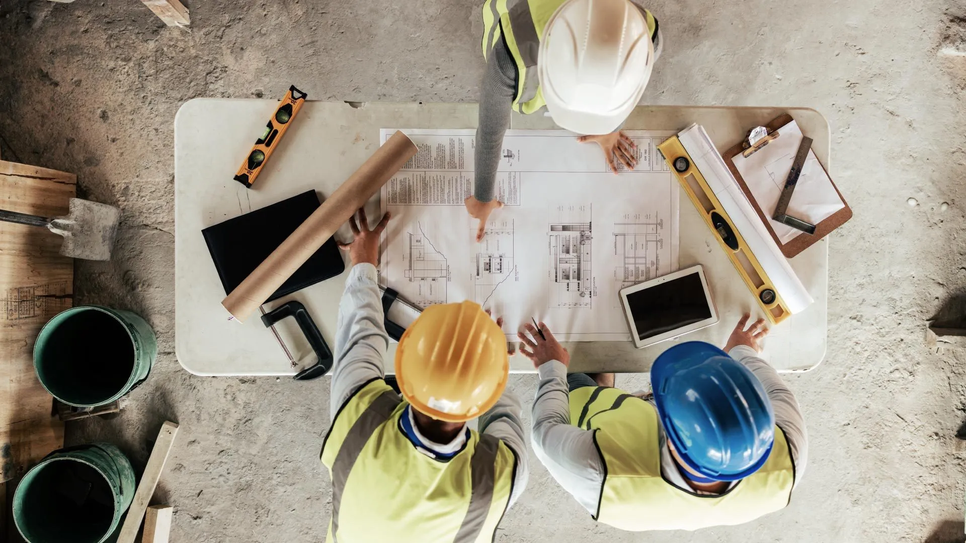 Three builders in hard hats reviewing construction drawings on a site table